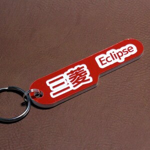 Könnte beinhalten: Roter Schlüsselanhänger mit einem silbernen Ring. Der Schlüsselanhänger trägt das Wort "Eclipse" und japanische Schriftzeichen in weiß.