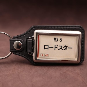 Puede incluir: Un llavero de cuero negro con un marco metálico rectangular y una inserción blanca. La inserción muestra el texto MX-5, el texto japonés para Roadster y un pequeño círculo rojo con las letras JP. Un anillo de metal plateado está unido al lateral.