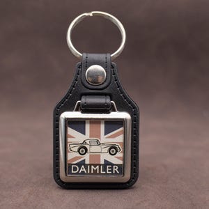 Puede incluir: Llavero de cuero negro con un marco de metal plateado. El marco presenta un diseño de la Union Jack y una silueta de coche blanco, con la palabra "DAIMLER" debajo. Un anillo plateado está unido en la parte superior.