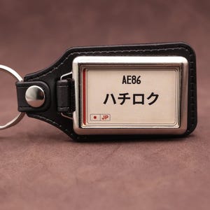 Pode incluir: Um porta-chaves de couro preto com uma moldura metálica retangular e uma inserção branca. A inserção apresenta o texto AE86, os caracteres japoneses ハチロク e um pequeno círculo vermelho ao lado das letras JP. Um anel de metal prateado está fixado na lateral.