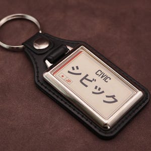 Honda Civic Japanese plate medallion keyring with black leatherette fob, chrome frame and cream insert on a brown suede background.