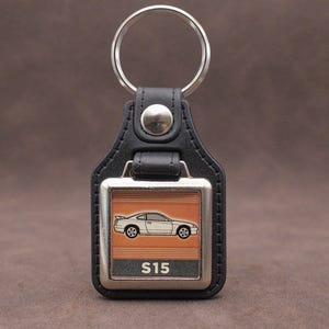 May include: Black leather keychain with a silver metal frame. The frame features a car silhouette over an orange background, with the text "S15" below. A silver key ring is attached.