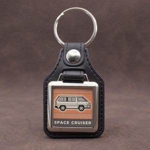 以下が含まれることがあります： 黒い革製のキーホルダー。銀色の金属フレームには、白い文字で「SPACE CRUISER」と書かれたバンのイラストが描かれています。キーホルダーには銀色のキーリングが付いています。