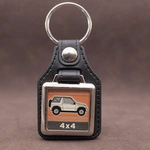 Op de afbeelding: Zwarte leren sleutelhanger met een zilverkleurig metalen frame. Het frame heeft een witte SUV-afbeelding op een oranje achtergrond, met de tekst "4x4" eronder. Een zilveren sleutelring is bevestigd.