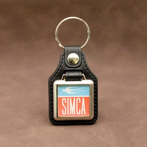 Può includere: Portachiavi in ​​pelle nera con un ciondolo quadrato argentato che presenta un uccello bianco e la parola "SIMCA" in lettere rosse su sfondo blu.