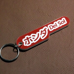 Könnte beinhalten: Roter Schlüsselanhänger mit einer weißen Aufschrift, die "Del Sol" und "オレンジ" in japanischen Schriftzeichen lautet.