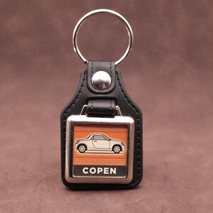 Peut inclure: Porte-clés en cuir noir avec un cadre en métal argenté. Le cadre présente une silhouette de voiture blanche sur fond orange, avec le mot "COPEN" en dessous. Un anneau argenté est attaché en haut.