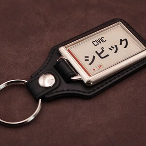 Honda Civic Japanese plate medallion keyring with black leatherette fob, chrome frame and cream insert on a brown suede background.
