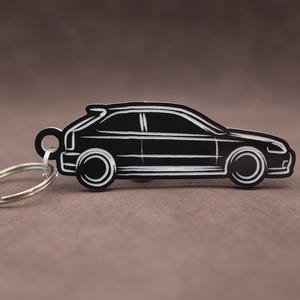 Peut inclure: Porte-clés en forme de voiture noire et blanche. Le porte-clés représente le profil d'une voiture à hayon avec des détails blancs sur les roues, les fenêtres et la carrosserie. Le porte-clés est attaché à un porte-clés argenté.