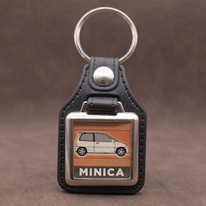 Op de afbeelding: Zwarte leren sleutelhanger met een zilverkleurige metalen plaat. De plaat toont een witte auto-illustratie en het woord "MINICA". De sleutelhanger heeft een zilveren ring.