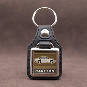 May include: Black leather keyring with a silver metal plate. The plate displays a car graphic and the name "CARLTON". The keyring has a silver ring and a button fastening.
