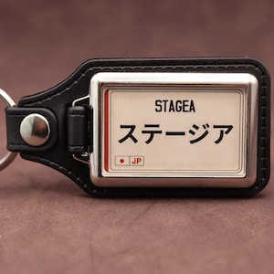 May include: A black leather keychain featuring a rectangular metal insert with a white background. The insert displays the text STAGEA in black, the Japanese characters ステージア, and a small red circle with the letters JP in a box.