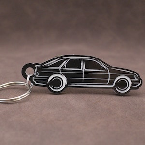 May include: A black car-shaped keyring with white detailing. The keyring depicts a classic car design, with wheels, windows, and a key ring. This car keyring is a stylish accessory for car enthusiasts.