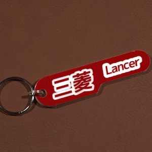 May include: Red keychain with a white metal ring. The keychain has the word "Lancer" and Japanese characters in white on a red background.