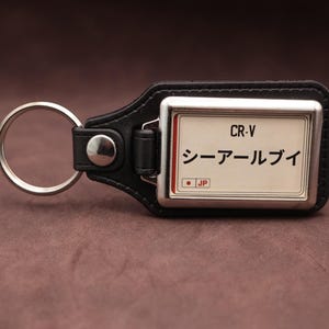 Puede incluir: Un llavero de cuero negro con una pieza metálica rectangular de cara blanca. La pieza muestra el texto CR-V, caracteres katakana japoneses y un pequeño punto rojo con las letras JP. Un anillo de metal plateado está unido al cuero.