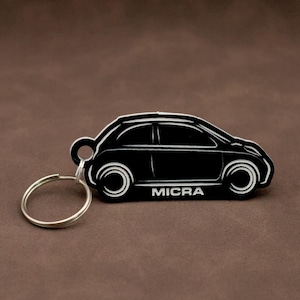 Könnte beinhalten: Schwarzer Autoschlüsselanhänger mit einem silbernen Schlüsselring. Der Schlüsselanhänger ist in Form eines Nissan Micra gestaltet und hat den Schriftzug "MICRA" auf der Seite.