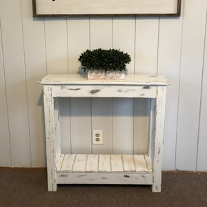 May include: White farmhouse style console table with a distressed finish. The table has a single shelf and is 30 inches tall. The table is in front of a white wall with a sign that says "farmhouse" in black lettering.