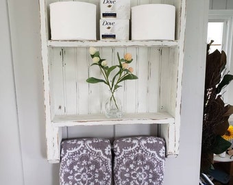 May include: White wooden shelf with two shelves and a towel rack. The top shelf has two rolls of toilet paper and two boxes of Dove soap. The middle shelf has a small vase with a sprig of flowers. The bottom towel rack has two gray towels with a white floral pattern.