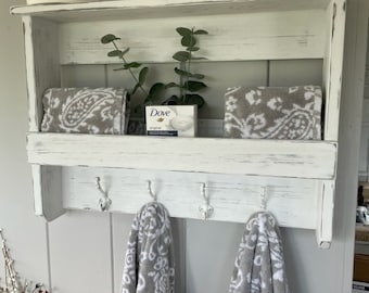 May include: White wooden bathroom shelf with two shelves and three hooks. The shelf has a distressed white finish and is decorated with a green candle, a plant, and towels. The word "Dove" is visible on a bar of soap.