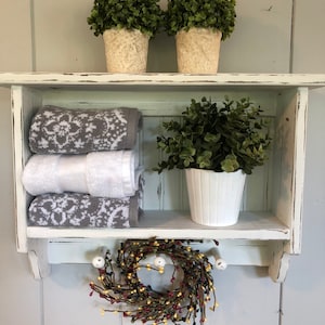 May include: White wooden shelf with two shelves and hooks on the bottom. The top shelf has two small potted plants with green leaves. The bottom shelf has two white towels, a potted plant with green leaves, and a small wreath with red, yellow, and black berries.