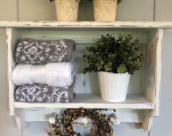May include: White wooden shelf with two shelves and hooks on the bottom. The top shelf has two small potted plants with green leaves. The bottom shelf has two white towels, a potted plant with green leaves, and a small wreath with red, yellow, and black berries.