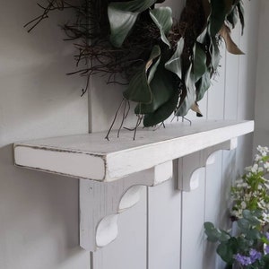 May include: White wooden shelf with decorative brackets. The shelf is mounted on a white wall.
