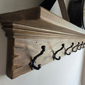 May include: A wooden shelf with a decorative trim and multiple black metal hooks. The shelf is stained a dark brown color and has a rustic appearance.