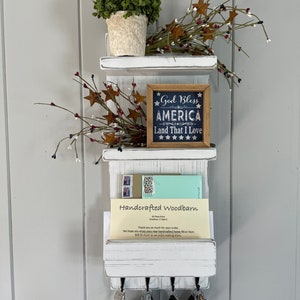 May include: White wooden key holder with a mail slot and three shelves. The top shelf has a small potted plant. The middle shelf has a sign that reads "God Bless America Land That I Love". The bottom shelf has a handwritten note that says "Handcrafted Woodbarn".