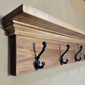 May include: A wooden shelf with a decorative trim and six black metal hooks. The shelf is stained a dark brown color.
