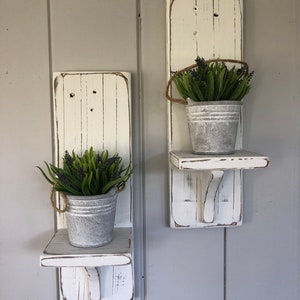 May include: Two white painted wooden wall shelves with a small metal bucket and faux greenery on each shelf. The shelves have a distressed look and are mounted on a gray wall.