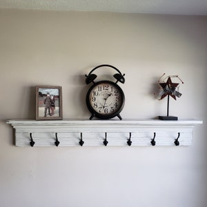 May include: A white painted wooden shelf with six black metal hooks. The shelf has a decorative molding around the top edge. A black alarm clock with a white face and black numbers is on the shelf. A framed photo and a red and white star decoration are also on the shelf.