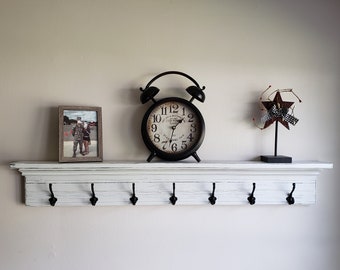 May include: A white painted wooden shelf with six black metal hooks. The shelf has a decorative molding around the top edge. A black alarm clock with a white face and black numbers is on the shelf. A framed photo and a red and white star decoration are also on the shelf.