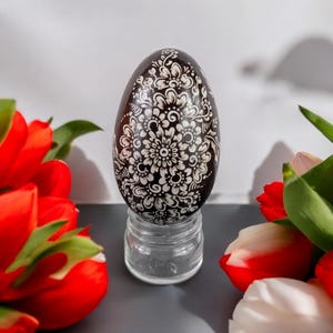 Op de afbeelding: Een bruin paasei met een wit bloemmotief wordt getoond op een transparante glazen standaard. Het ei wordt omringd door rode tulpen.