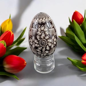 May include: A brown Easter egg with a white floral design is displayed on a clear glass stand. The egg is surrounded by red and yellow tulips.