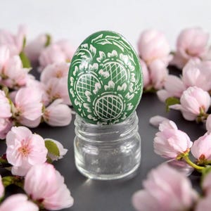 Può includere: Un uovo di Pasqua verde con un motivo floreale bianco è esposto su un barattolo di vetro trasparente. L'uovo è circondato da fiori di ciliegio rosa.