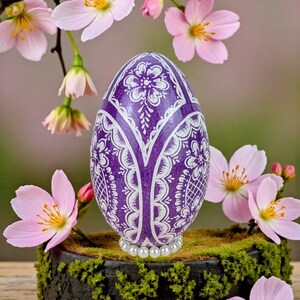 Pode incluir: Um ovo de Páscoa roxo com padrões florais brancos e uma base de pérolas. O ovo é exibido sobre uma cama de musgo verde, rodeado por flores de cerejeira rosa. O design do ovo apresenta detalhes brancos intrincados.