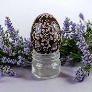 Könnte beinhalten: Ein braunes Osterei mit einem weißen Blumenmuster wird auf einem klaren Glasständer präsentiert. Das Ei ist von Lavendelblüten umgeben.