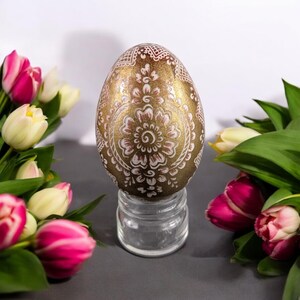 Pode incluir: Um ovo de Páscoa dourado com um desenho floral branco e rosa é exibido em um suporte de vidro transparente. O ovo é cercado por tulipas rosa e brancas.