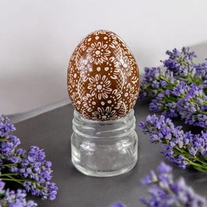 Könnte beinhalten: Ein braunes Osterei mit weißem Blumenmuster wird auf einem klaren Glasständer präsentiert. Das Ei ist von Lavendelblüten umgeben.