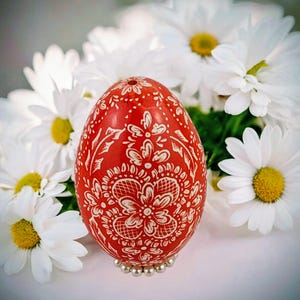 Peut inclure: Un œuf de Pâques rouge vif orné de motifs floraux blancs complexes. L'œuf est présenté sur un lit de petites perles argentées, avec en arrière-plan des marguerites blanches à cœur jaune. Le motif floral est symétrique.