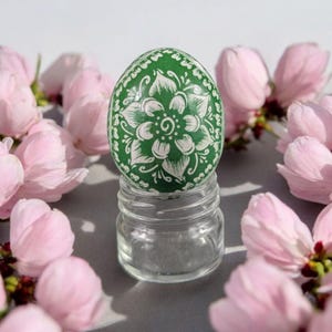 Può includere: Un uovo di Pasqua verde con un motivo floreale bianco, appoggiato su un piccolo barattolo di vetro. L'uovo è circondato da delicati fiori rosa, creando un'estetica primaverile. Il design dell'uovo presenta un fiore centrale e motivi dettagliati.
