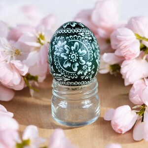 May include: A green Easter egg with a white floral design sits on a clear glass jar. The egg is surrounded by pink blossoms.