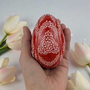 Könnte beinhalten: Ein rotes dekoratives Ei mit weißen Blumenmustern. Das Ei wird in einer Hand gehalten, mit weißen Tulpen im Hintergrund. Das Design des Eies zeigt komplizierte weiße Blumenmotive, die einen auffälligen Kontrast erzeugen.