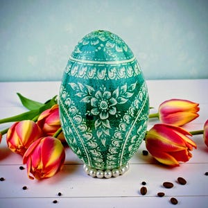 Op de afbeelding: Een turquoise paasei met witte bloempatronen en een parelbasis. Het ei is omringd door rode en gele tulpen en koffiebonen, wat een lentethema creëert. Het ontwerp van het ei bevat ingewikkelde bloemen- en krulwerk.