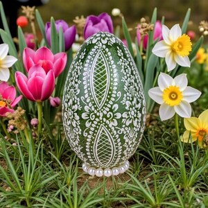 Könnte beinhalten: Ein dekoratives grünes Osterei mit weißen floralen und geometrischen Mustern. Das Ei ist mit einer Perlenbasis verziert und inmitten von leuchtend rosa Tulpen, weißen Narzissen und grünem Gras platziert, was ein Frühlingsmotiv erzeugt.
