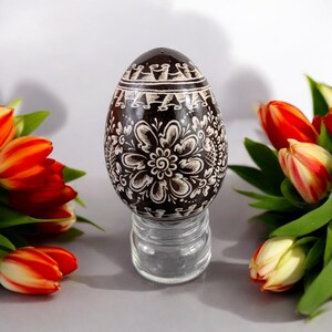 May include: A brown Easter egg with a white floral design is displayed on a clear glass stand. The egg is surrounded by tulips with red and white petals.
