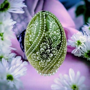 Op de afbeelding: Een groen, versierd ei met witte bloempatronen. Het ei is versierd met ingewikkelde ontwerpen en rust op een basis van kleine parels. Witte bloemen en een paarse achtergrond omringen het ei.