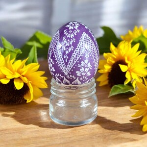 Könnte beinhalten: Ein violettes Osterei mit einem weißen Spitzenmuster wird auf einem Glasgefäß präsentiert. Das Ei ist von gelben Sonnenblumen umgeben.
