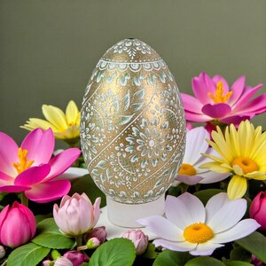 Op de afbeelding: Een decoratief gouden ei met ingewikkelde witte en lichtgroene bloempatronen, staand op een witte basis. Het ei is omgeven door levendige roze, gele en witte bloemen met groene bladeren, wat een feestelijke en kleurrijke weergave creëert.