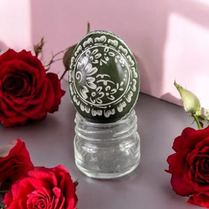 Op de afbeelding: Een olijfgroen ei, versierd met witte bloemmotieven, rust op een klein glazen potje. Rode rozen omringen het ei, tegen een roze en grijze achtergrond.
